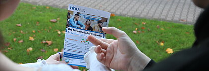 Auf dem Foto ist der Flyer vom Buddy-Programm zu sehen. Zwei Hände zeigen auf dem Flyer