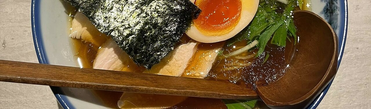 Eine Nudelsuppe mit Ei, Seetang, Kräutern und Fleisch mit einem länglichen Holzlöffel 