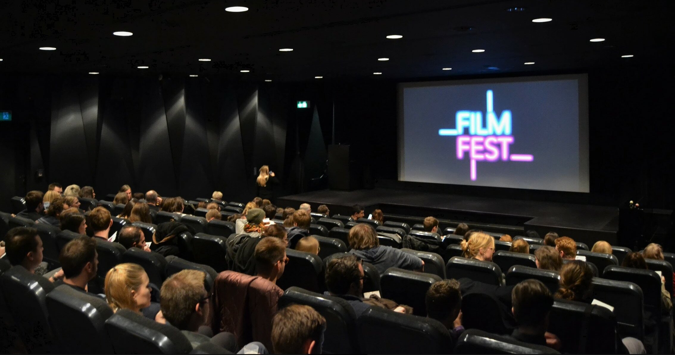 Zu sehen ist ein Kinosaal gefüllt mit Menschen, auf der Leinwand ist "Filmfest Düsseldorf" angezeigt.