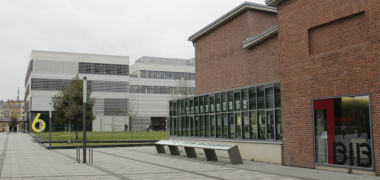 Zu sehen ist der Eingang der Bibliothek. Liks sind die Fenster des Erinnerungsorts im Bild. Eine Infotafel erstreckt sich über die gesammte Länge der Fensterfront. Im Hintergrund ist der Campus der HSD zu sehen.