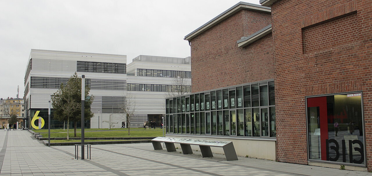 Außenansicht des Erinnerungsorts und Bibliothekeingangs (Foto: Eva Marschall) Zu sehen ist der Eingang der Bibliothek. Liks sind die Fenster des Erinnerungsorts im Bild. Eine Infotafel erstreckt sich über die gesammte Länge der Fensterfront. Im Hintergrund ist der Campus der HSD zu sehen.