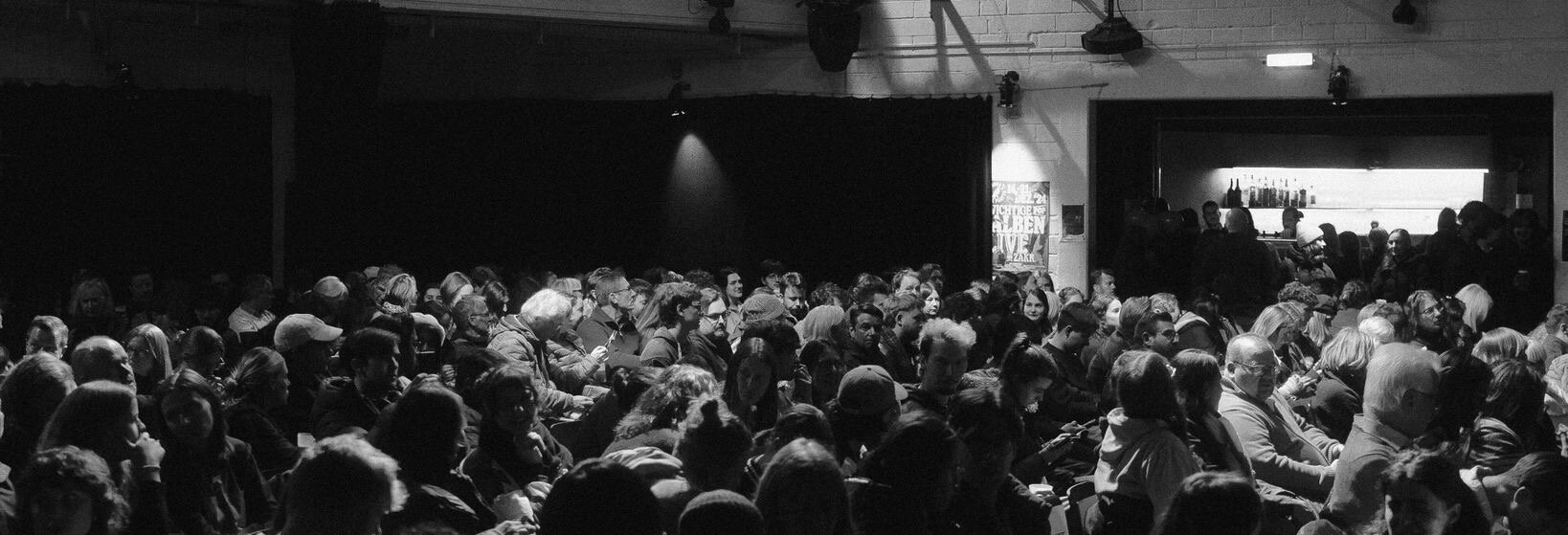 Auf dem Foto ist ein Publikum in schwarz-weiß zu sehen, was vor einer Bühne steht.