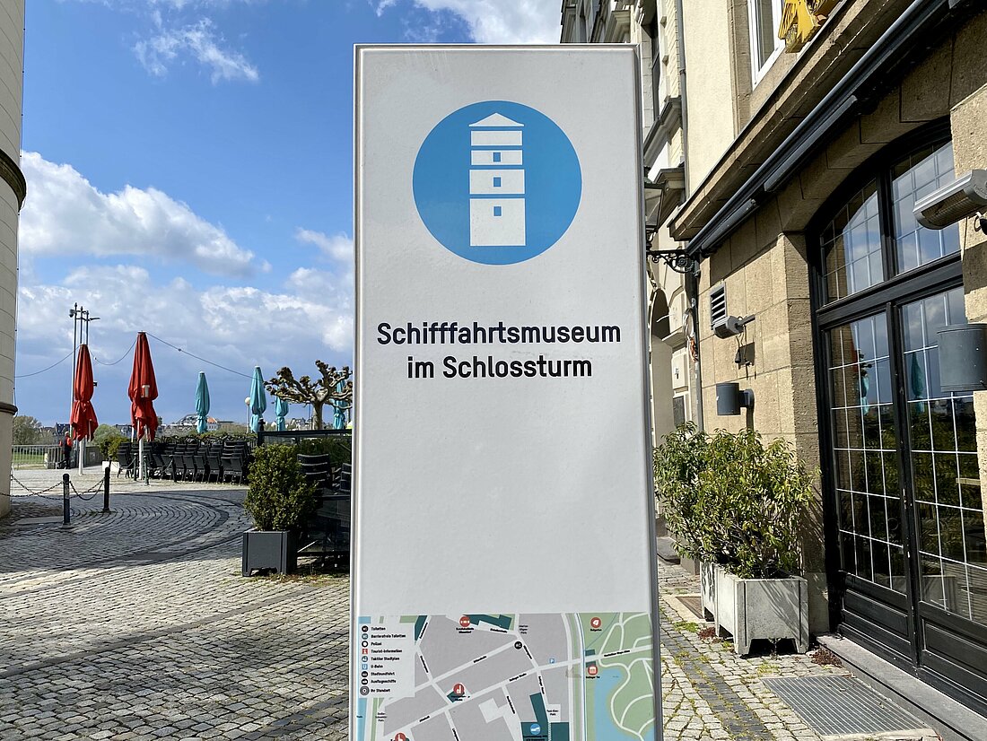 Das Informationsschild des Museums neben dem Schlossturm und Restaurants im Hintergrund.