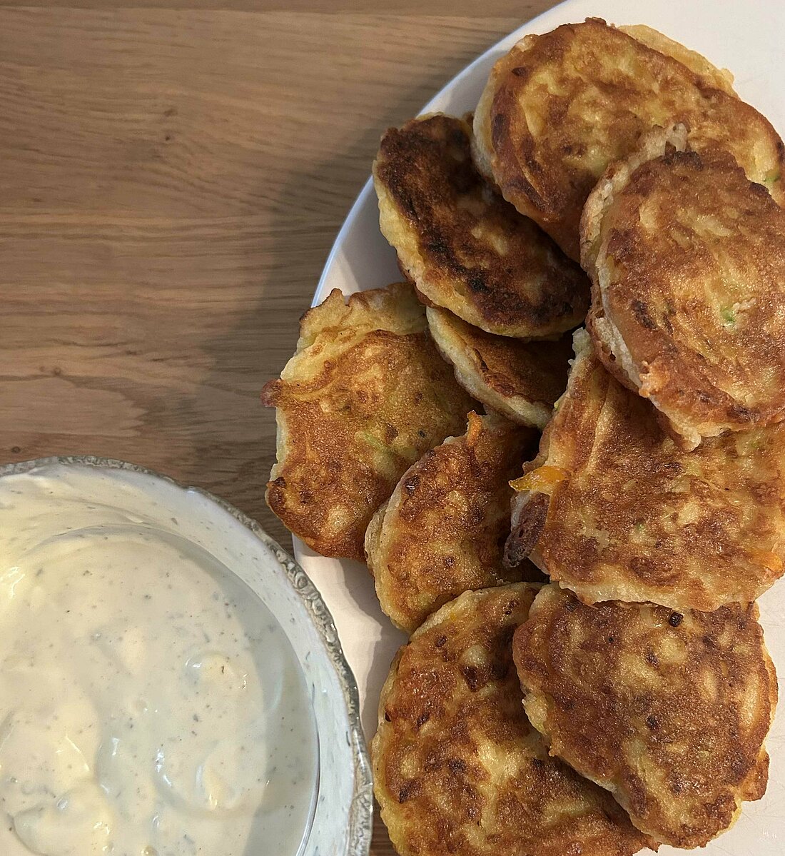 Auf dem Bild sieht man Zucchini Pancakes mit einem Dip serviert. Beides steht auf einem Tisch mit einem Teller (Foto: Lena Sorgalla)
