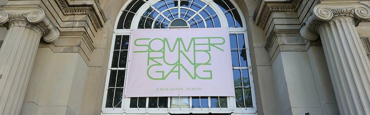 Auf dem Foto sieht man den Eingang der Kunstakademie und ein pinkes Banner mit der grünen Aufschrift "Sommerrundgang 2026"