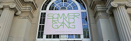Auf dem Foto sieht man den Eingang der Kunstakademie und ein pinkes Banner mit der grünen Aufschrift "Sommerrundgang 2026"