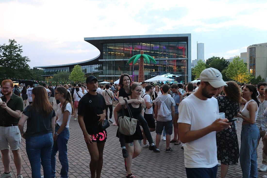 Mehrere Studierende sind auf dem Platz vor der Bibliothek. Eine Studentin wird von einer anderen Studentin huckepack getragen.