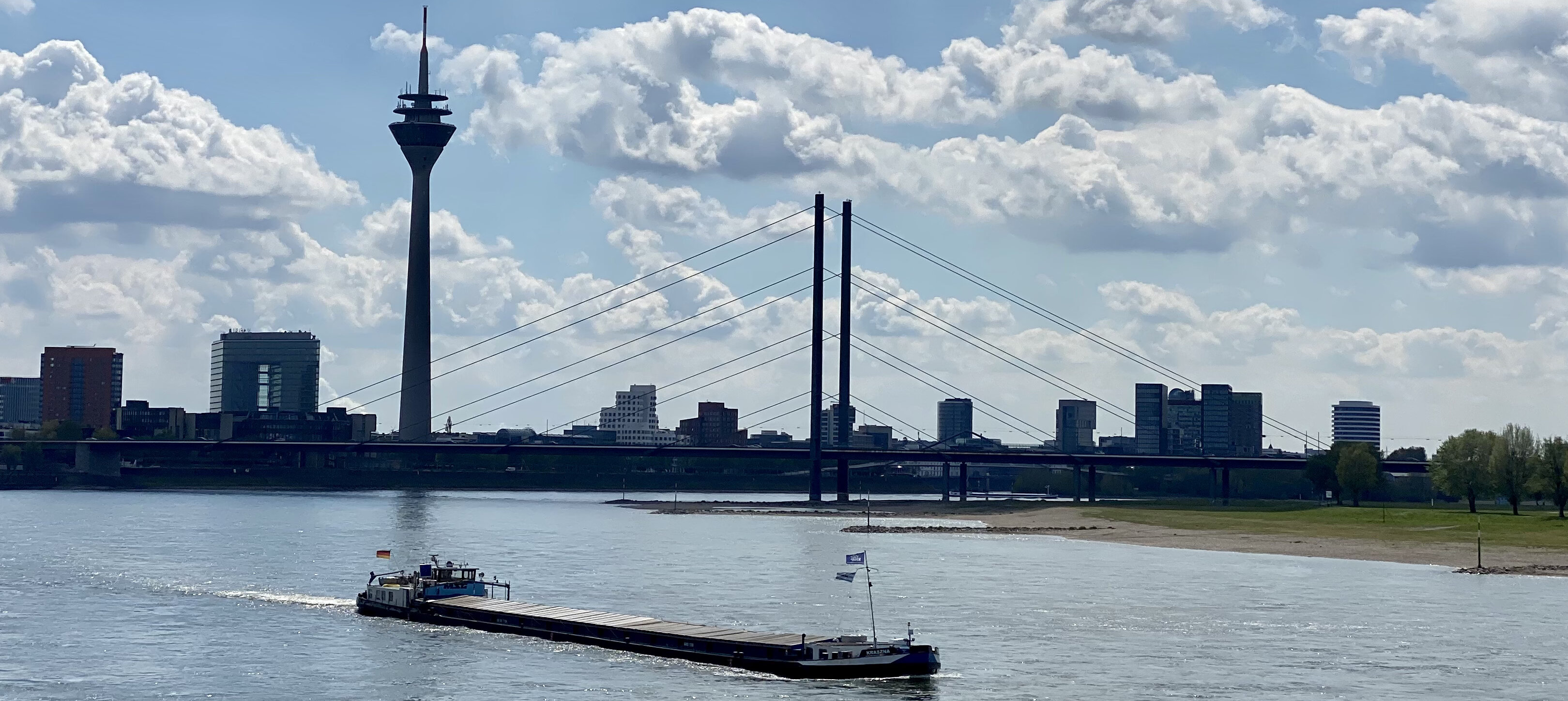 Auf dem Foto sieht man die Düsseldorfer Skyline bei gutem Wetter mit einem Boot im Vordergrund auf dem Rhein
