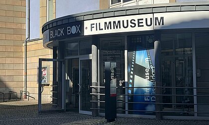 Auf diesem Bild sieht man den Eingang des Filmmuseums. Die Tür steht offen und darüber ist das Museum sowie das hauseigene Kino die Blackbox ausgeschildert. 