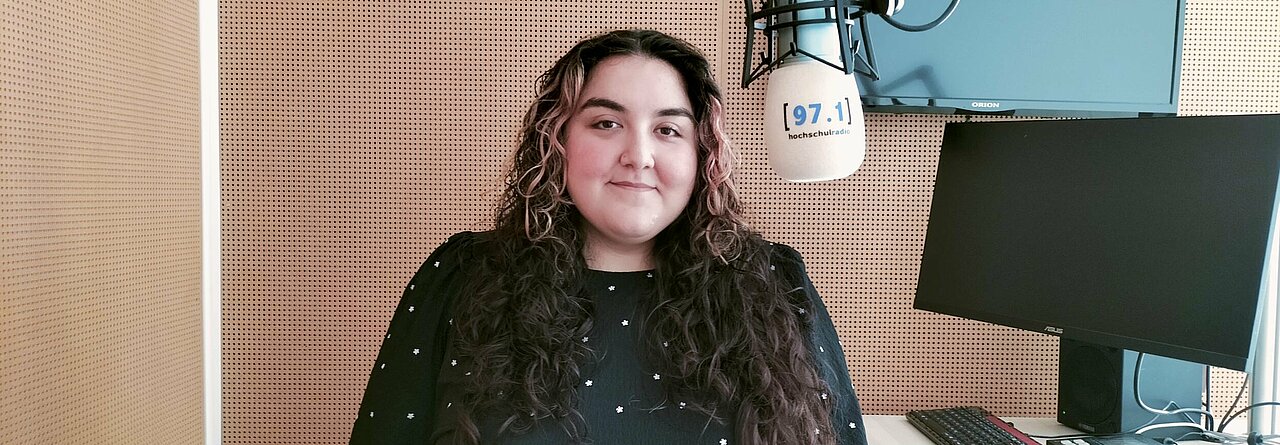 Auf dem Foto ist Elif Benli zu sehen. Sie steht neben einem Mikrofon und lächelt in die Kamera.