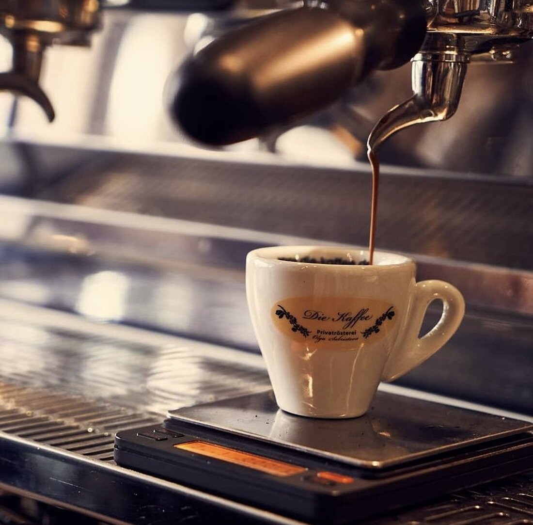 Auf dem Bild sieht man eine Espresso Tasse, mit einem Aufdruck der Rösterei, die unter einer Siebträgermaschine steht. Aus dem Siebträger läuft der frische Espresso direkt in die kleine Tasse.
