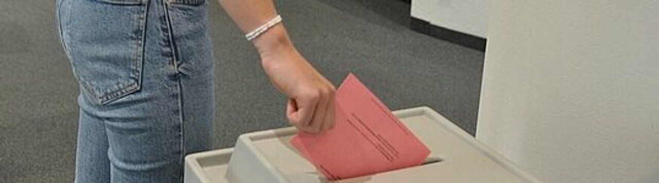 Auf dem Foto ist eine Wahlurne zu sehen. Jemand wirft gerade einen roten Wahlzettel ein.