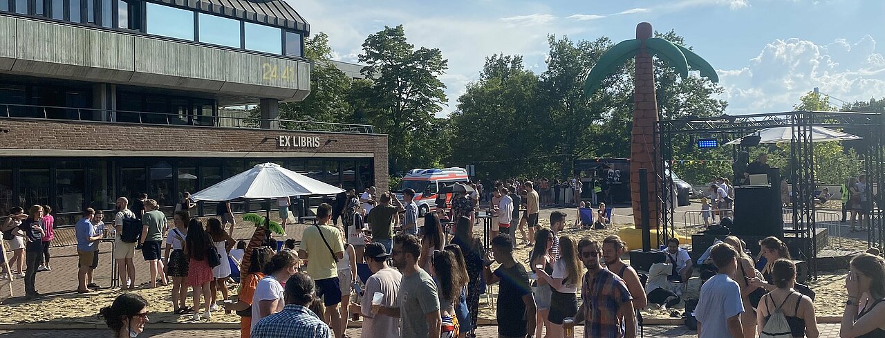 Die Summer Beach Party vor der Bibliothek (Foto: Jacqueline Kowalczyk) Man sieht mitten auf dem Campus der Heinrich-Heine-Universität eine Sandflächen mit Schirm, einer kleinen Luftpalme darunter auf der linken Seite und rechts eine große Luftpalme und weiter rechts eine kleine Bühne für den DJ. Auf der Sandfläche, dahinter und davor ist eine Menschenmenge versammelt.