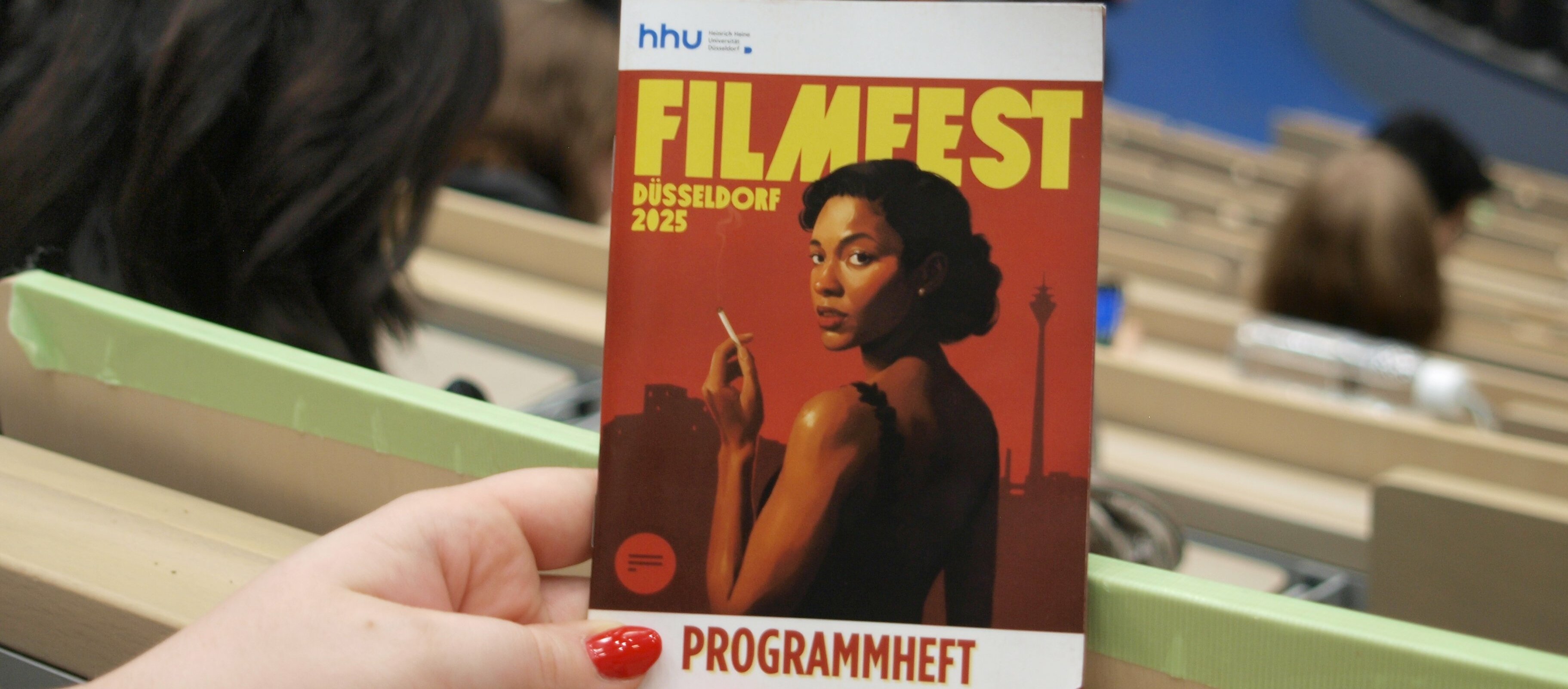 Auf dem Foto sieht man das das Programmheft vom Flmfest. Vorne ist das Plakat drauf. Eine Frau hält es in der Hand im Hörsaal 3A.