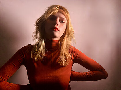 Die Musikerin Kira Hummen (Foto: Karla Lee) Kira Hummen trägt einen roten Rollkragenpullover und schaut in die Kamera. Sie hat ihre Arme in die Seite gestützt und steht vor einem rosa Hintergrund.