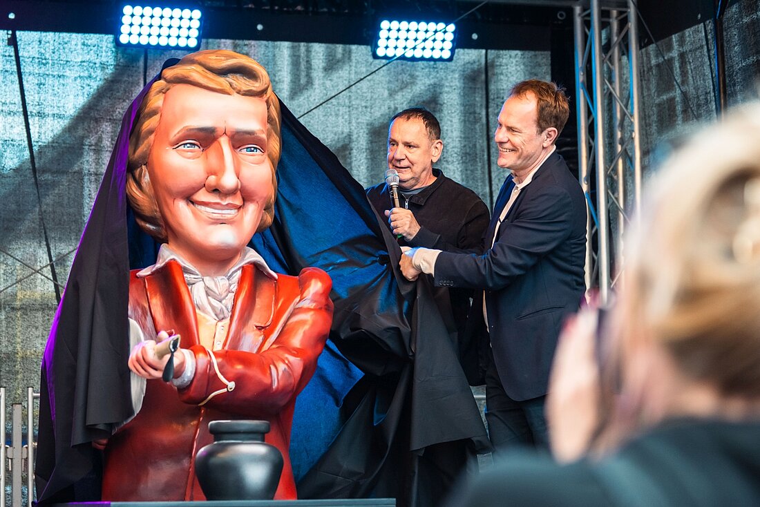 Auf dem Foto sind Oberbürgermeister Stephan Keller und Jacques Tilly zu sehen. Sie stehen neben der neuen Statue von Heinrich Heine und enthüllen sie gerade.