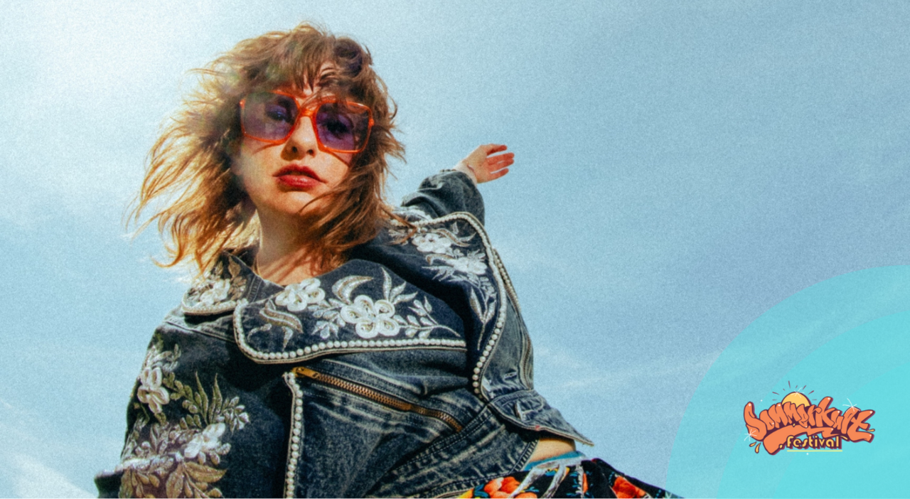Mola dreht sich unter blauem Himmel und streckt die Arme weit aus. Sie trägt eine Sonnenbrille und eine Jeansjacke mit Stickereien.