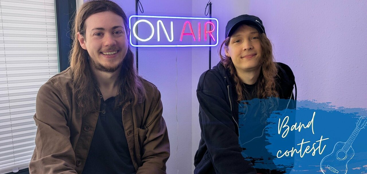 Sören und Juna sitzenen nebeneinander vor einem leuchtenden On Air Schild. Sie lächeln in die Kamera.
