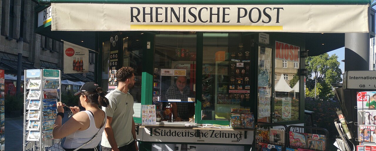 Man sieht ein Kiosk von aussen; der Besitzer schaut durch seine Verkaufsscheibe direkt in die Kamera. Davor schauen sich zwei Kunden das Sortiment an.