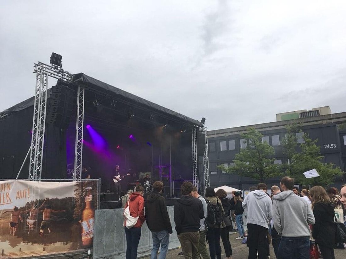 Man sieht eine Bühne mit Musiker:innen. Davor steht Studierende.