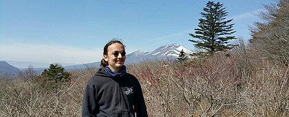 Man sieht einen Studierenden der HHU vor einer japanischen Berglandschaft. Die Sonne scheint ihm ihn ins Gesicht und er lächelt zufrieden.