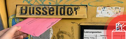 Auf dem Foto ist ein gelber Briefkasten zu sehen. Jemand wirft gerade eine roten Wahlbrief ein.