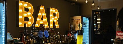 Maria_Bartheke Auf dem Foto sieht man eine Bar, das Wort Bar geschrieben mit bunten Lichtern und hängend an der Wand. Vorne sieht man ein paar Getränke