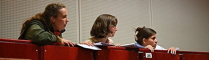Auf dem Bild sind die drei Schauspielerinnen zu sehen wie sie sich hinter Hörsaalstühlen verstecken und gespannt zum rechten Bildrand schauen (Foto: Andreas Meske)