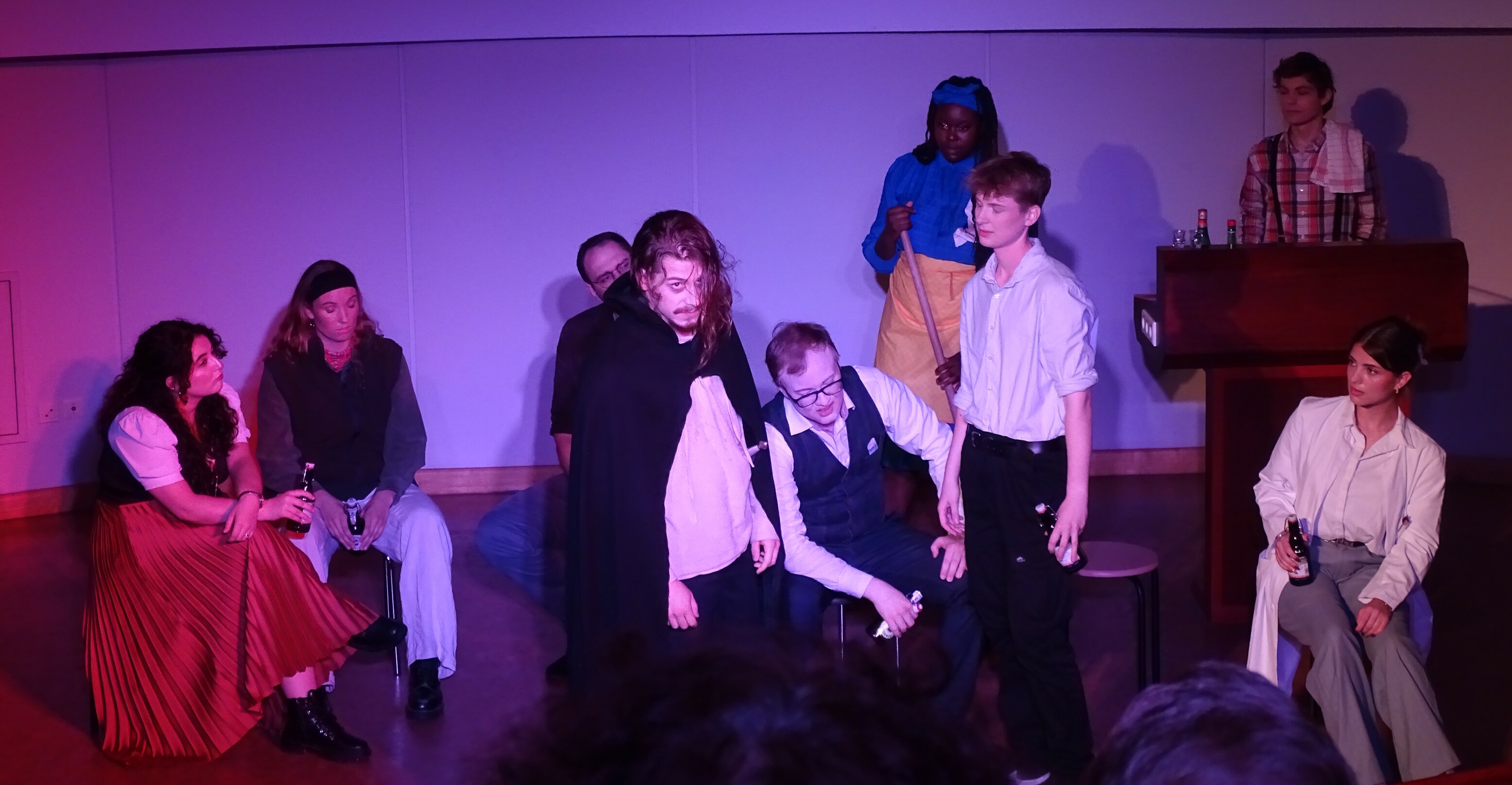 Auf dem Bild ist ein Großteil des Ensembles zu sehen. Die Bühne soll hier eine Kneipe darstellen und Ratti steht im Mittelpunkt der Aufmerksamkeit.