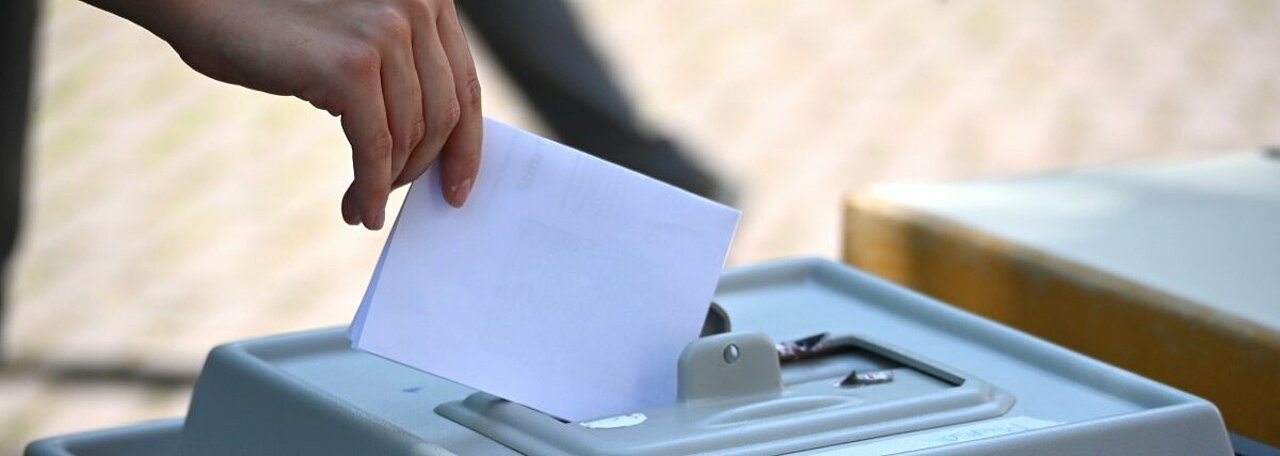 Auf dem Foto ist eine Wahlurne zu sehen. Jemand wirft gerade einen Wahlzettel in die Urne.