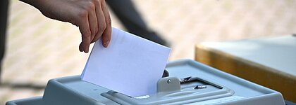 Auf dem Foto ist eine Wahlurne zu sehen. Jemand wirft gerade einen Wahlzettel in die Urne.
