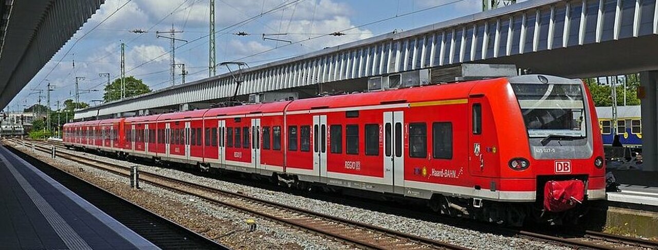 Zu sehen ist ein roter Zug, der auf Gleisen vor einem Bahnsteig steht. 
