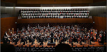 Auf dem Bild sieht man den Unichor und das Uniorchester in Aktion auf einer Bühne.