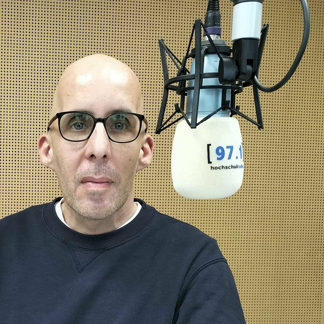Benedikt Walter steht im Studio von hochschulradio düsseldorf vor einem Mikrofon. Er blickt direkt in die Kamera und lächelt.