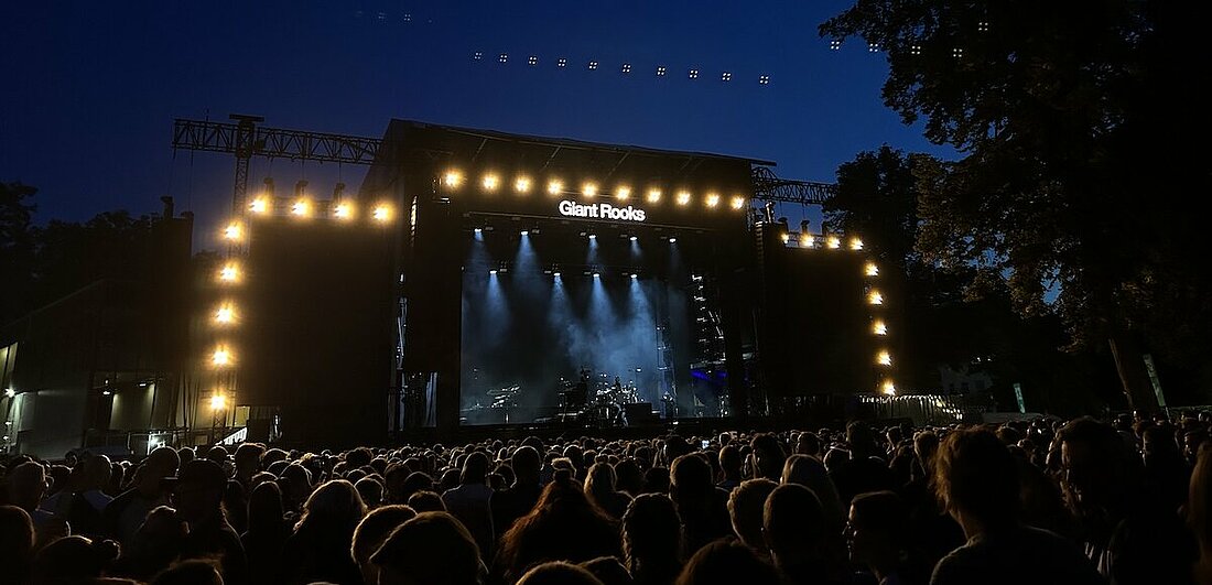 Man sieht die noch leere Bühne, die mit Leuchtstrahlern umrahmt wird. Am oberen Rand in der Mitte steht „Giant Rooks“. Vor der Bühne steht das Publikum.