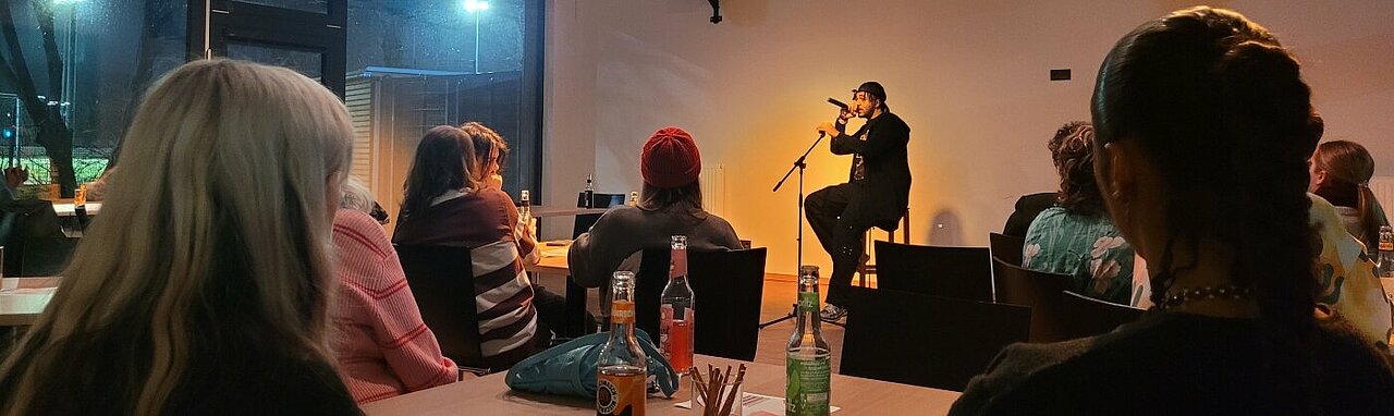 Auf dem Bild sieht man Leute in dem Cafe Jugendring in Düsseldorf sitzen. Sie hören dem Rapper CHAMP zu.