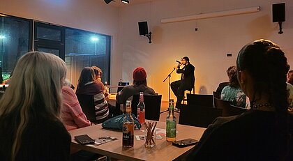 Auf dem Bild sieht man Leute in dem Cafe Jugendring in Düsseldorf sitzen. Sie hören dem Rapper CHAMP zu.