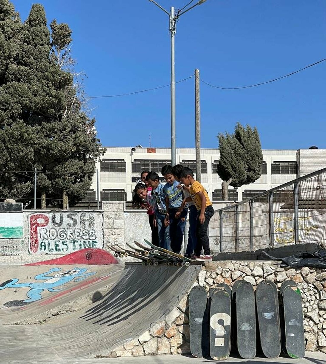 Sechs Kinder stehen dicht nebeneinander auf einer Rampe. Ihre Skateboards sind abfahrbereit auf der Kante platziert.