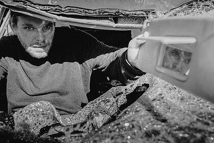 Schwarz-Weiß-Aufnahme eines Mannes, der auf dem Fahrersitz eines kaputten Autos sitzt und durch die zerstörte Frontscheibe in die Kamera schaut. In der Hand hält er den abgebrochenen Rückspiegel des Autos.
