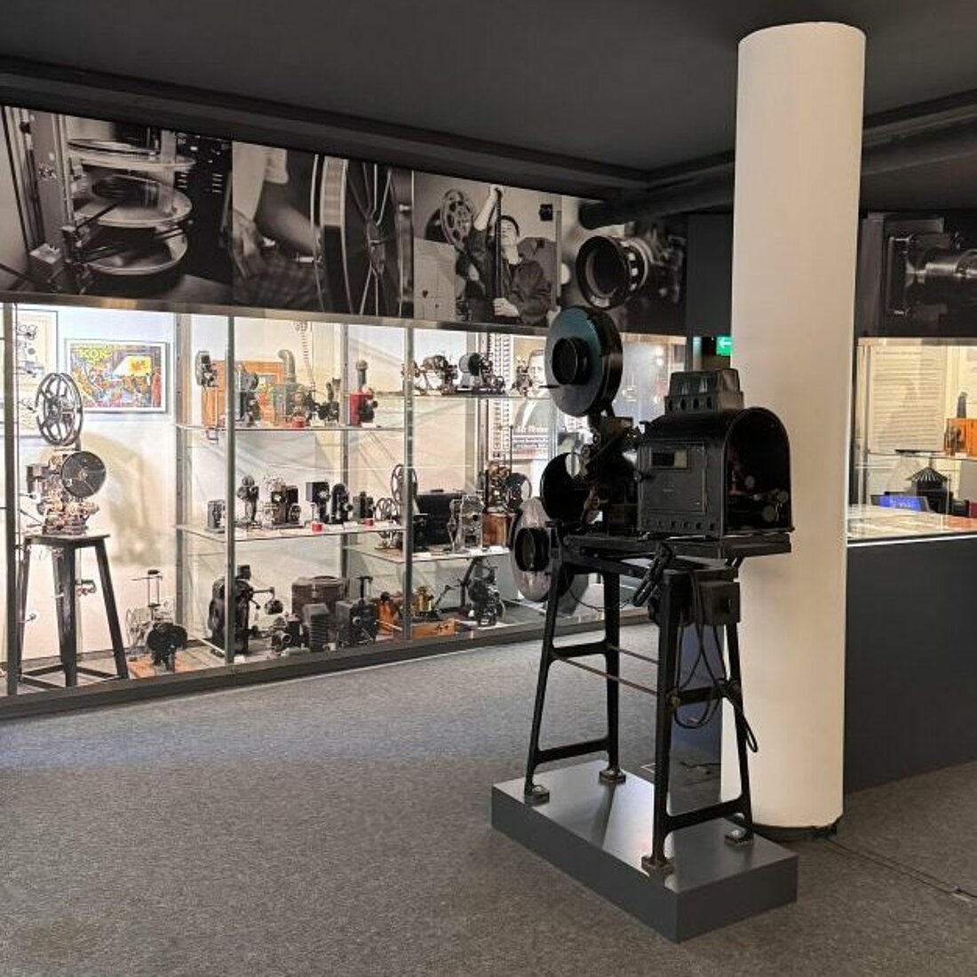 Auf dem Bild sieht man eine große Filmkamera in der Mitte eines Raumes. Glasvitrinen mit kleineren Kameras und Apparaten sind an den Wänden plaziert.