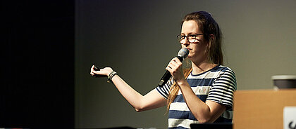 Anna Hülkenberg auf der Bühne bei einem Slam. (Foto: Klangblende) Anna Hülkenberg auf der Bühne bei einem Slam. Sie trägt einem gestreiftes T-Shirt und hält ein Mikrofon in der Hand.