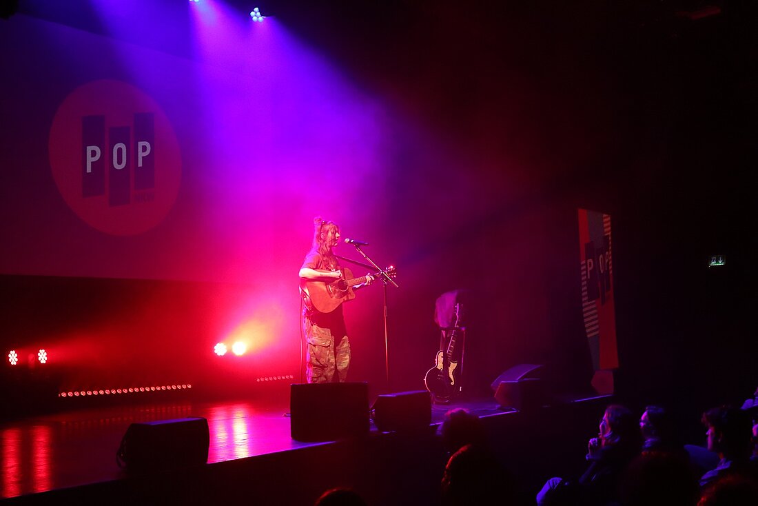 Zu sehen ist die Sängerin Philine Sonny während ihres Auftritts. Sie spielt Gitarre, im Hintergrund ist rotes und blaues Licht zu sehen.