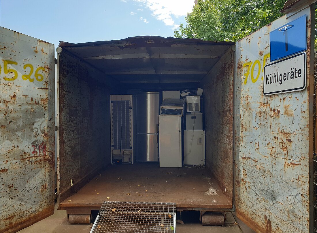 Man schaut von außen in einen geöffneten Schiffscontainer, es gibt einen starken Kontrast zwischen wolkenfreiem Himmel und dem dunklen Innern des rostigen Containers. Im Container sieht man alte Kühlschränke und Container gestapelt.