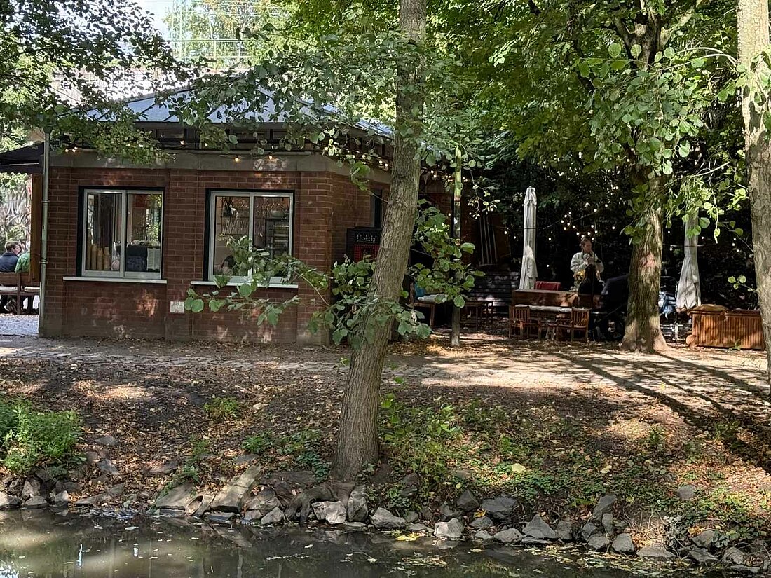 Man sieht ein kleines Gebäude, dass im Schatten der Bäume steht und sich direkt an einem Bach befindet 