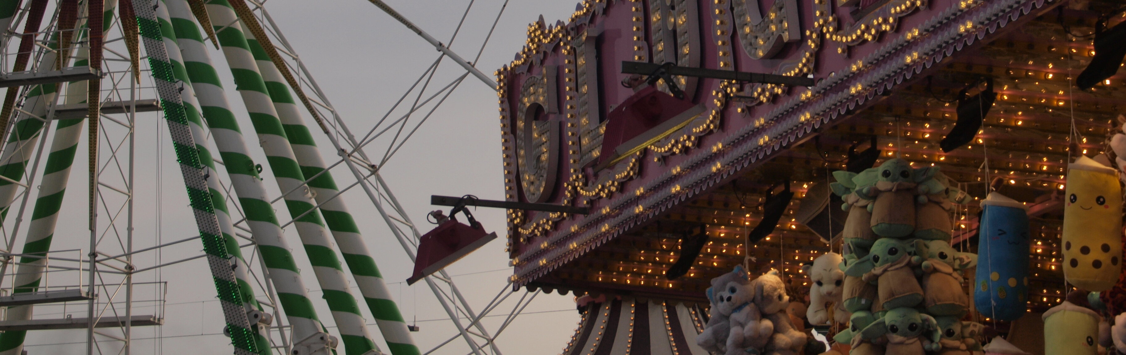 Auf dem Foto sieht man rechts einen Kirmesstand an dem Kuscheltiere hängen und links den Ausschnitt eines Riesenrads.