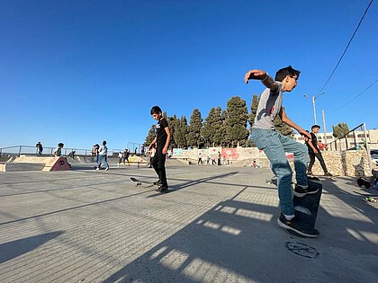 Man sieht den Skatepark unter blauem Himmel mit ca. 20 Personen, vor der Kamera übt ein Jugendlicher einen Trick auf seinem Board.
