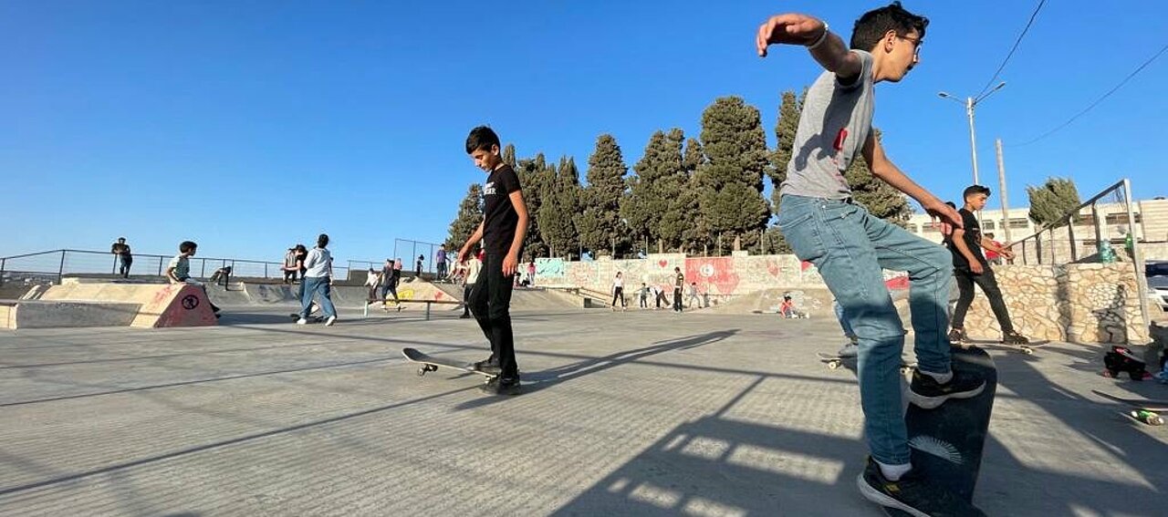 Man sieht den Skatepark unter blauem Himmel mit ca. 20 Personen, vor der Kamera übt ein Jugendlicher einen Trick auf seinem Board.