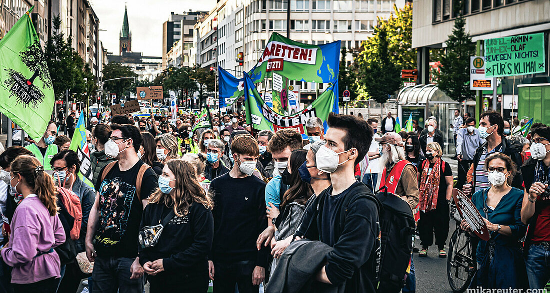 Auf dem Bild sieht man einen Demozug von Fridayforfuture in Düsseldorf. Im Vordergrund stehen junge Menschen, die medizinische Masken tragen und gespannt in die Ferne gucken. Im Hintergrund sieht man viele weitere Menschen aller  Altersgruppen, die Plakate, Banner und Fahnen beschriftet  mit Forderungen nach mehr Klimaschutz bei sich tragen.