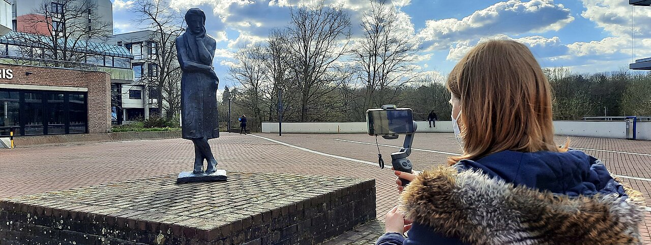 Eine CampusTV-Redakteurin steht vor der Heinrich-Heine-Statue auf dem Campus und filmt mit ihrem Smartphone. 