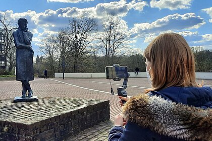 Eine CampusTV-Redakteurin steht vor der Heinrich-Heine-Statue auf dem Campus und filmt mit ihrem Smartphone. 